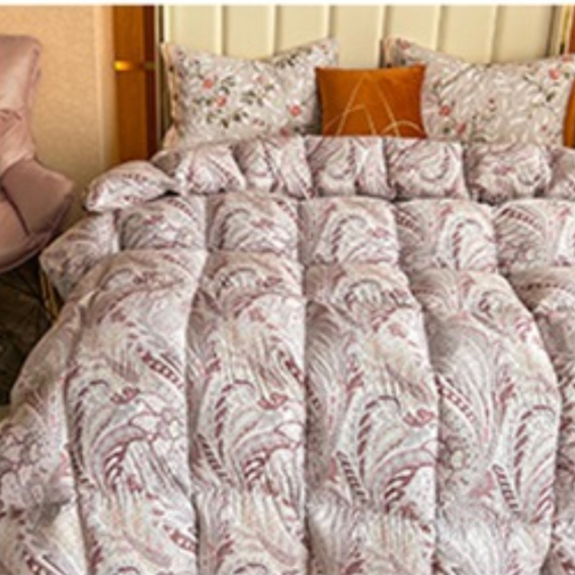 A close-up of a bed made with a patterned quilted comforter and matching pillows in a bedroom setting. The comforter has a dusty rose and beige botanical leaf pattern, with two floral pillows and one solid orange accent pillow.