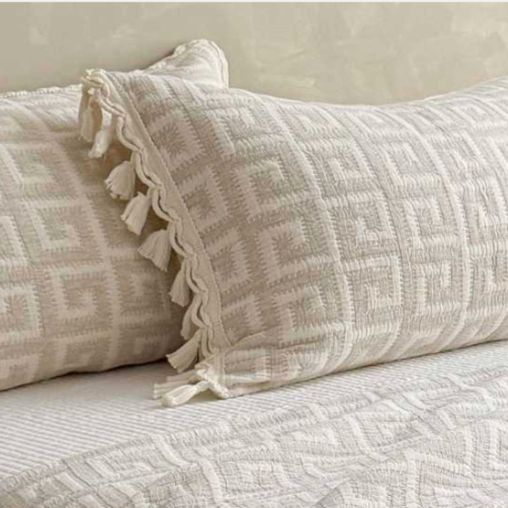 A close-up of textured cream and beige bedding with a geometric pattern, featuring a decorative lumbar pillow with tassels and matching pillows on a bed.