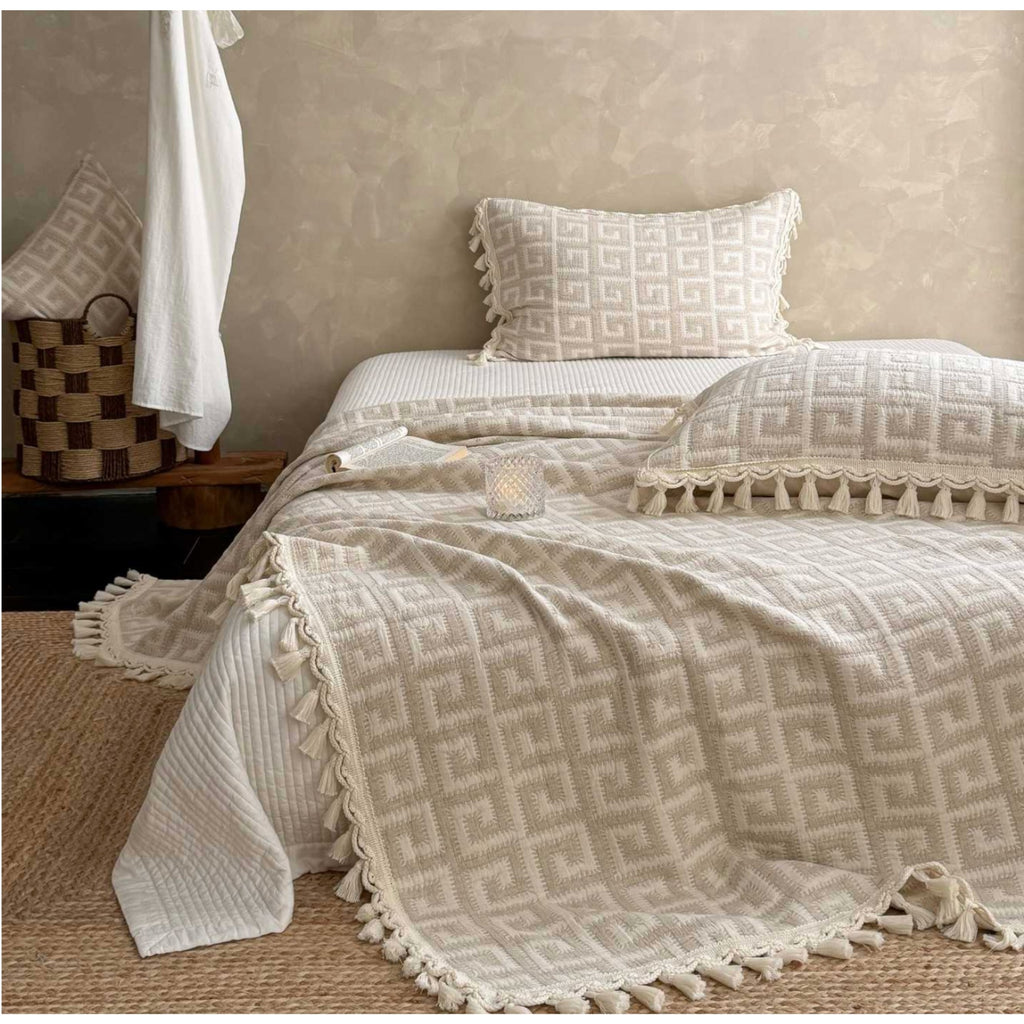 A beige and off-white textured throw blanket with tassels and a geometric pattern is styled on a bed, accompanied by matching pillows and a white hanging towel in a rustic bedroom setting.