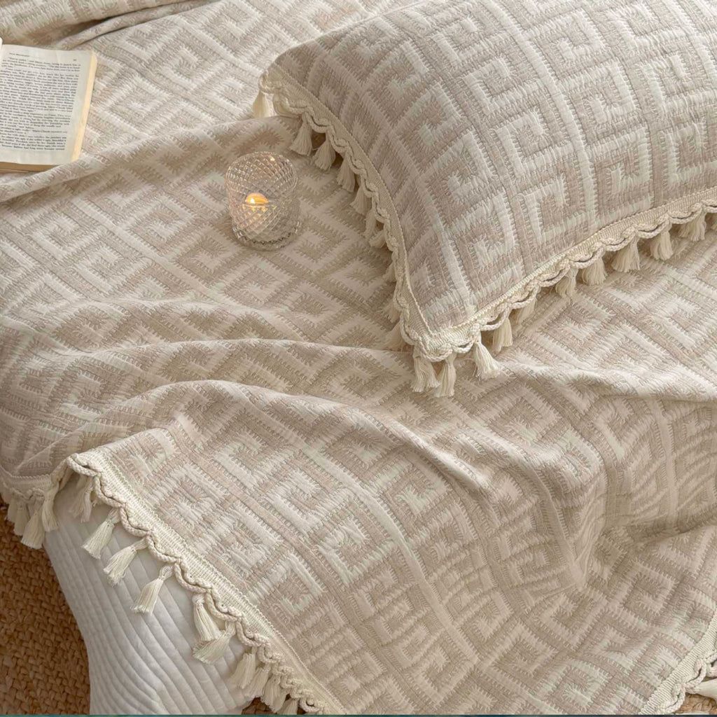 A close-up of a beige and cream textured throw blanket and matching pillowcase with a geometric pattern and tassel trim, styled on a bed with a small lit candle and an open book.