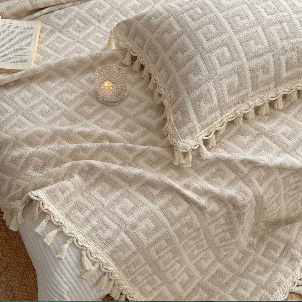 A close-up of a beige and cream textured throw blanket and matching pillowcase with a geometric pattern and tassel trim, styled on a bed with a small lit candle and an open book.