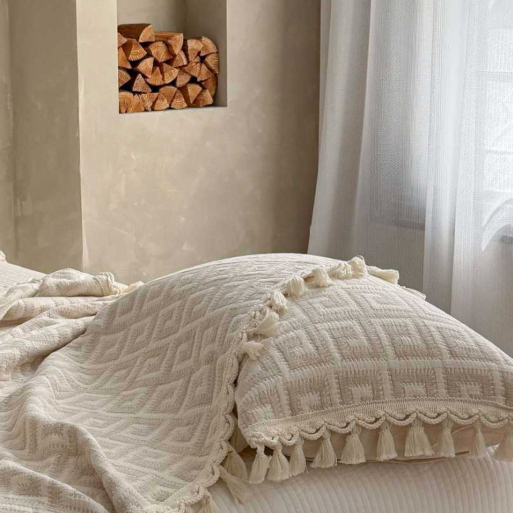 Cream-colored textured throw blanket and matching pillowcase with tassels, styled on a bed in a warm, neutral-toned bedroom setting.