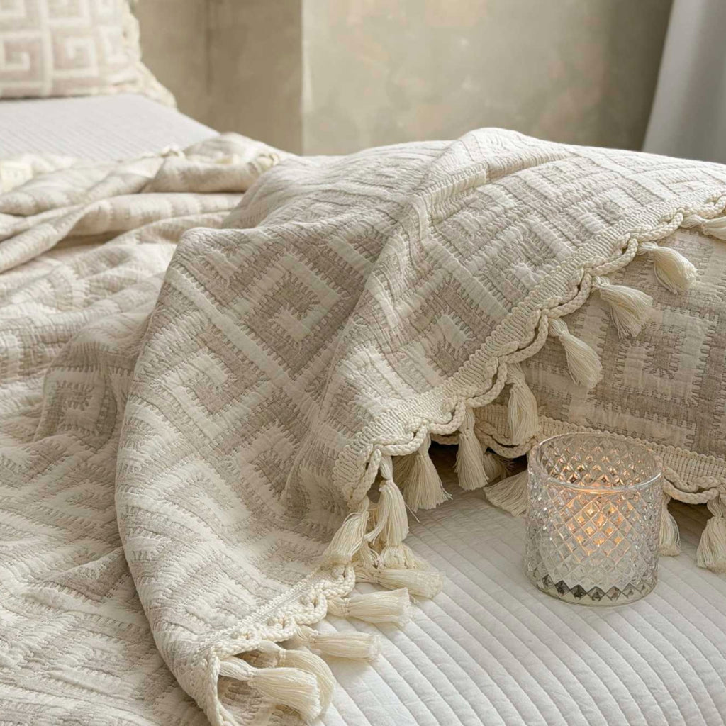 A cream-colored textured throw blanket with geometric patterns and tassels draped over a bed, next to a lit glass candle holder.
