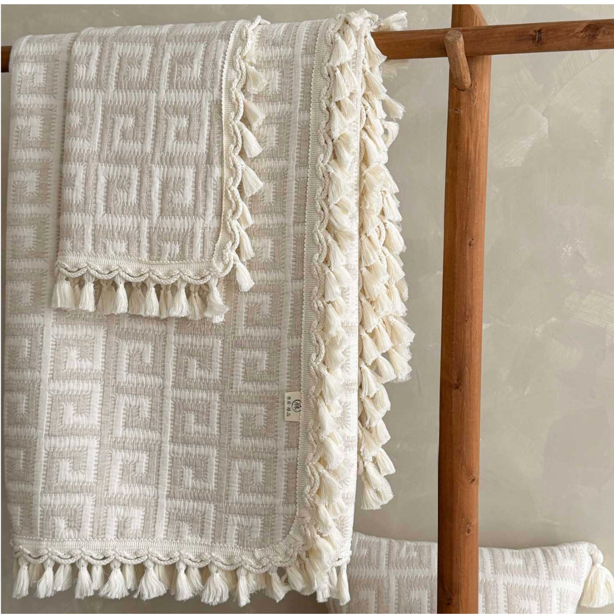 An off-white textured throw blanket with a geometric pattern and tassels, draped over a wooden rack.