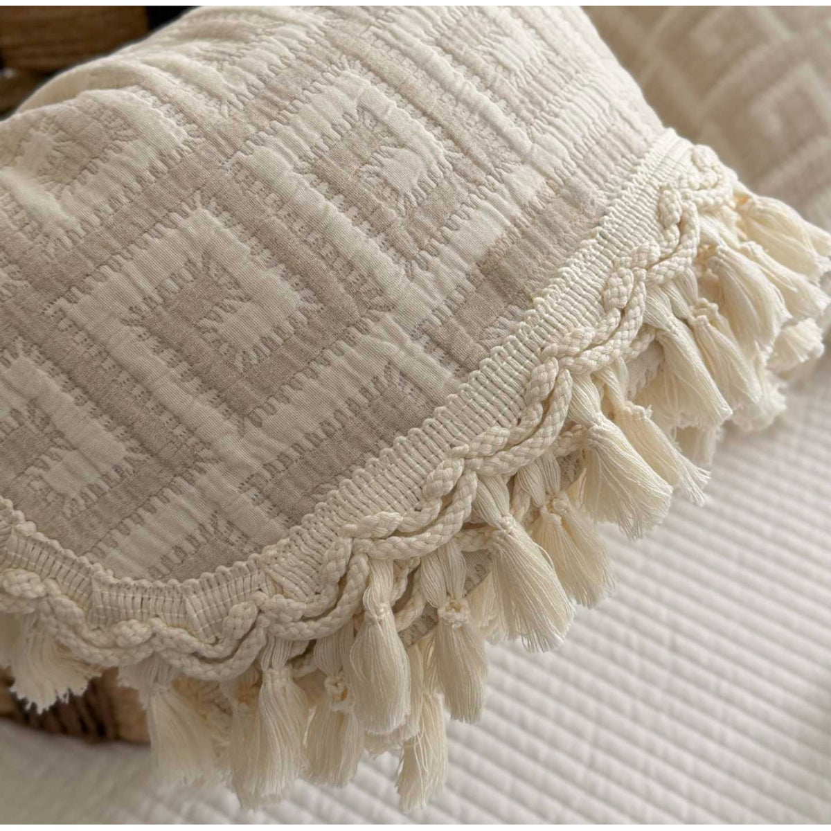 Close-up of a cream-colored textured throw pillow with a geometric woven pattern and a braided tassel trim, resting on a matching ribbed quilted surface.