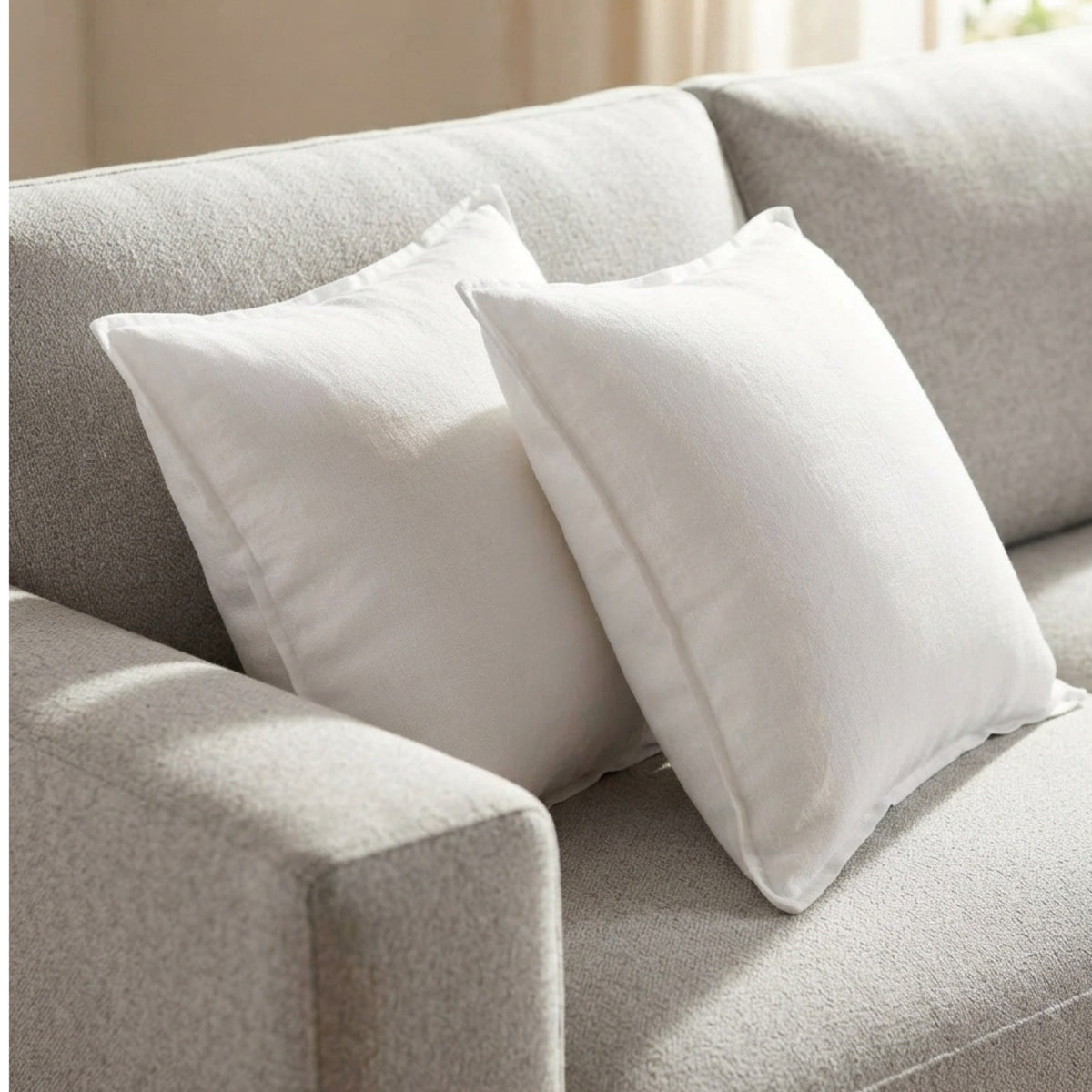 Two white square throw pillows resting on a light gray textured sofa in a softly lit interior setting.