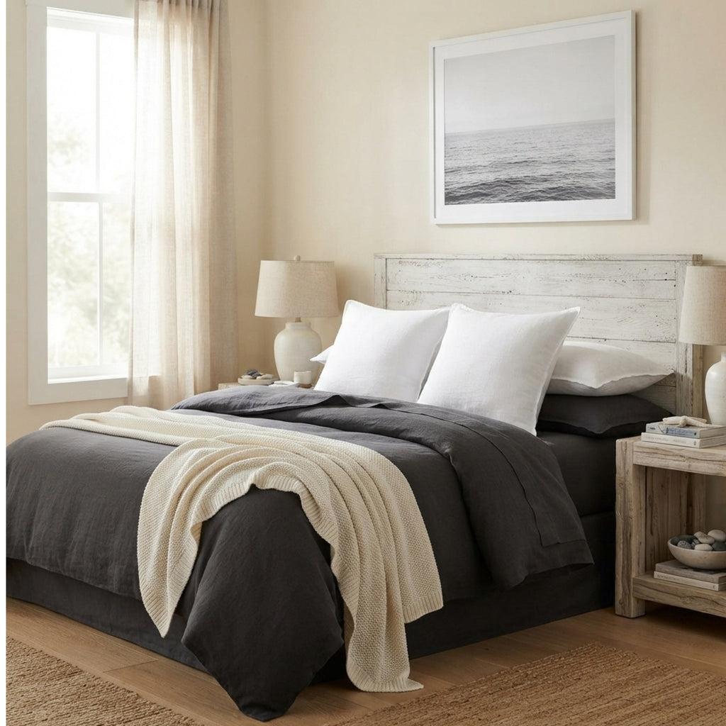 A neatly made bed in a softly lit bedroom, dressed in dark charcoal bedding with white pillows and a cream throw blanket draped over the foot of the bed. The room includes a rustic white wood headboard, matching nightstands, table lamps, a window with sheer curtains, and framed wall art.