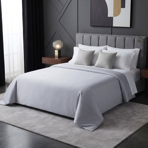 A neatly made bed dressed in light gray bedding with matching gray and white pillows, set in a modern bedroom with a gray upholstered headboard, dark walls, a side table with a lamp, and a light rug.