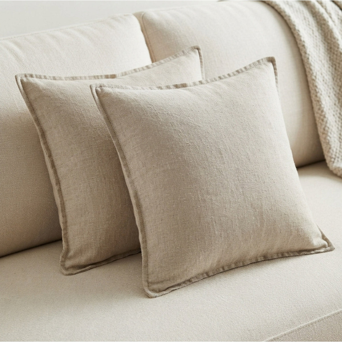 Two square beige linen pillows with a linen-like texture resting on a matching beige sofa, with a textured cream throw blanket draped over the back.