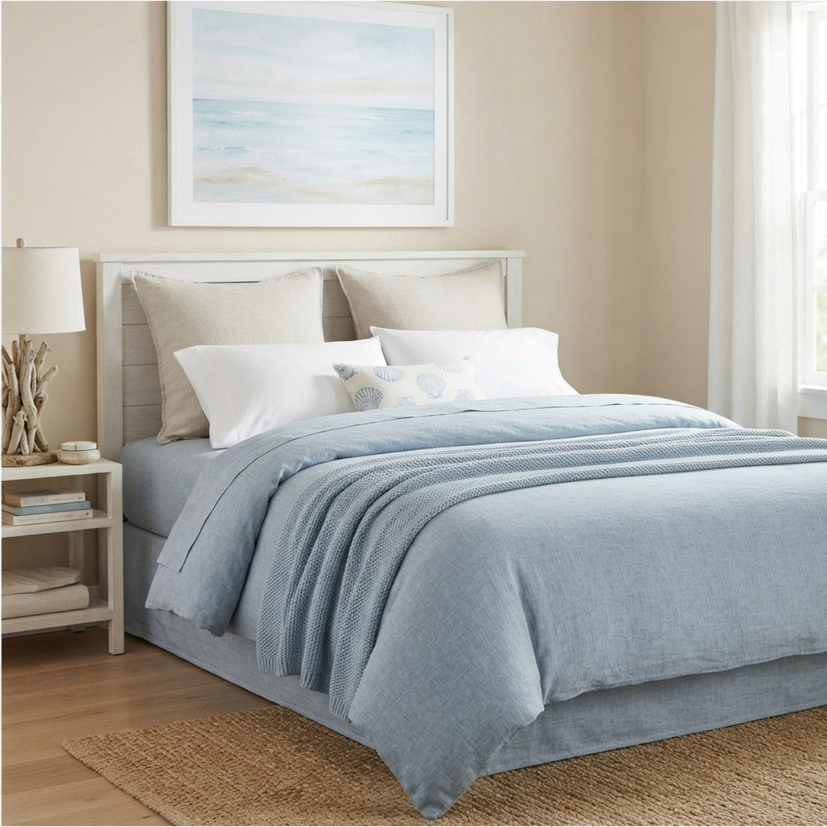 A neatly made bed in a softly styled bedroom, dressed with light blue bedding and white pillows, with a matching light wood headboard and bedside table visible.