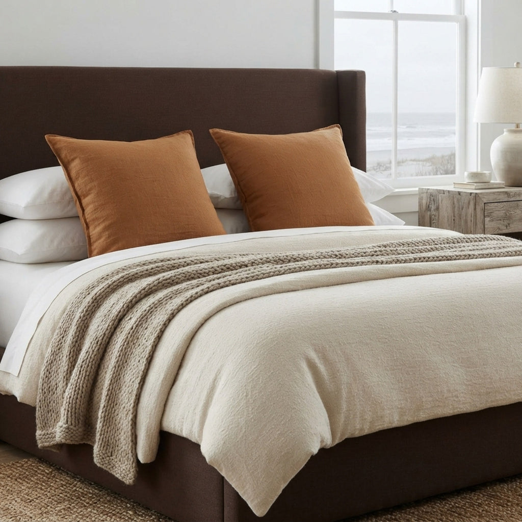 A styled bed in a bright bedroom featuring a dark brown upholstered bed frame, white bedding, two rust-orange accent pillows, and a textured beige throw blanket.