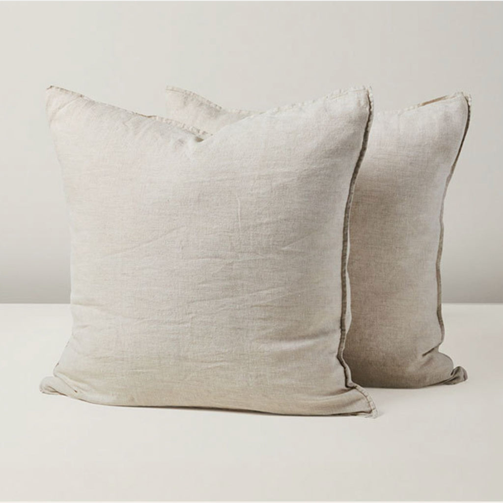 Two square throw pillows in a light beige linen fabric, with one pillow in front and another slightly behind it, set against a neutral off-white background.