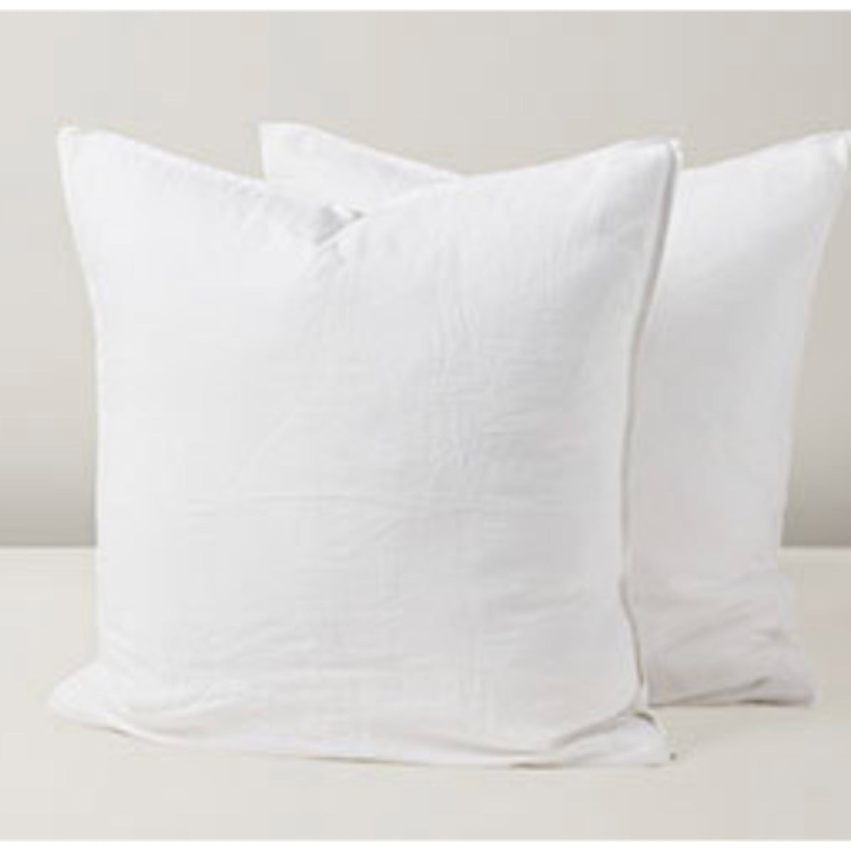 Two plain white square throw pillows with a textured fabric, arranged on a light beige surface against a neutral beige background.