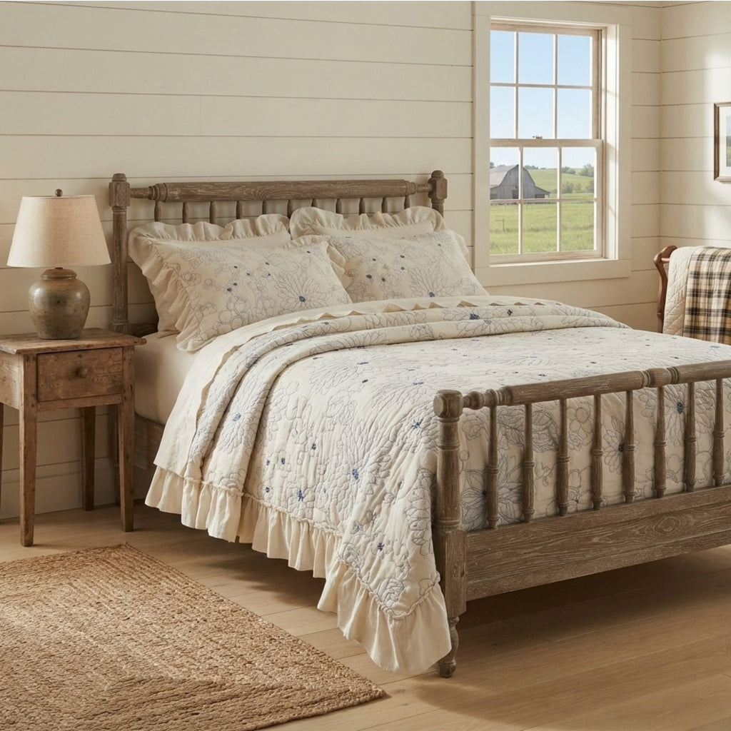 A rustic wooden bed frame dressed in a cream quilted bedding set with blue floral embroidery and ruffled edges, styled in a bright, shiplap-walled bedroom with a nightstand, lamp, and window view.