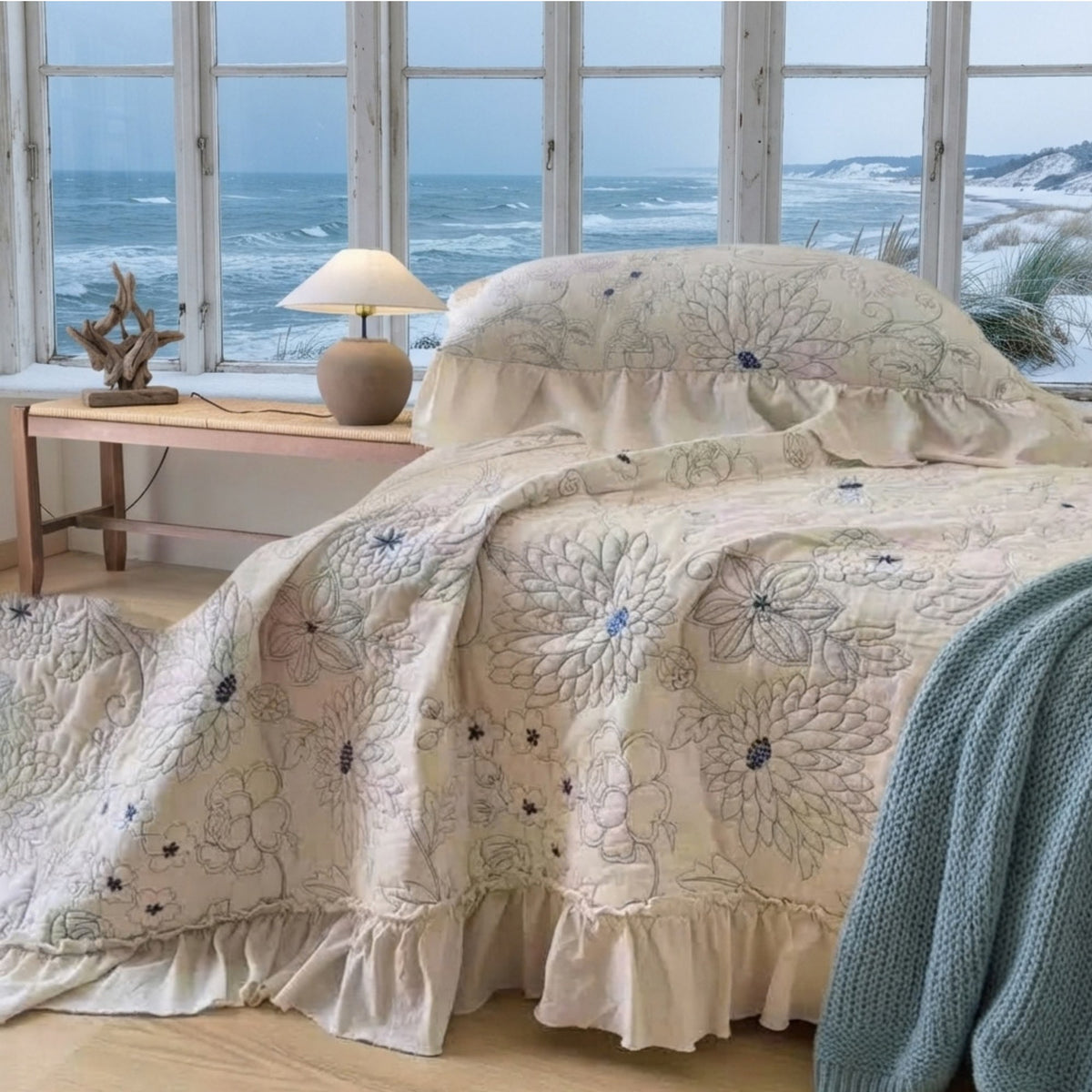 A bed dressed in a cream-colored quilted cover with large floral embroidery and ruffled edges, styled in a bright bedroom with large windows overlooking the ocean.