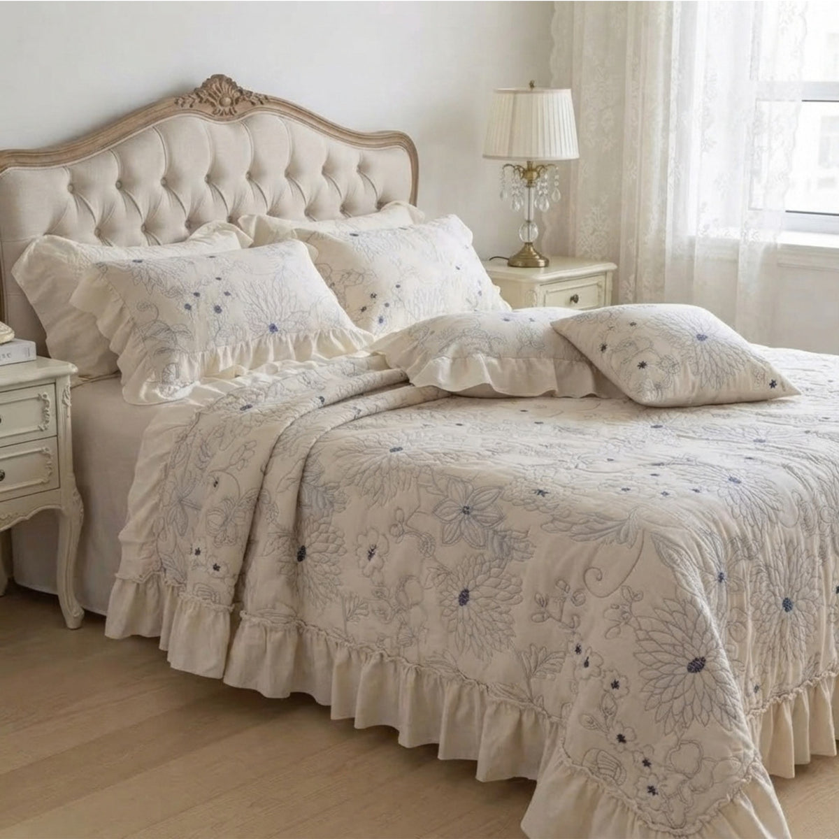 A cream-colored bedding set with blue floral embroidery and ruffled details, styled on a bed with a tufted headboard in a bright, classic bedroom.
