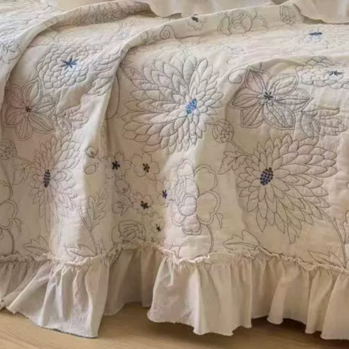 A close-up of a quilted bedspread with a ruffled skirt, featuring large floral embroidery in muted blue and gray on an off-white background.