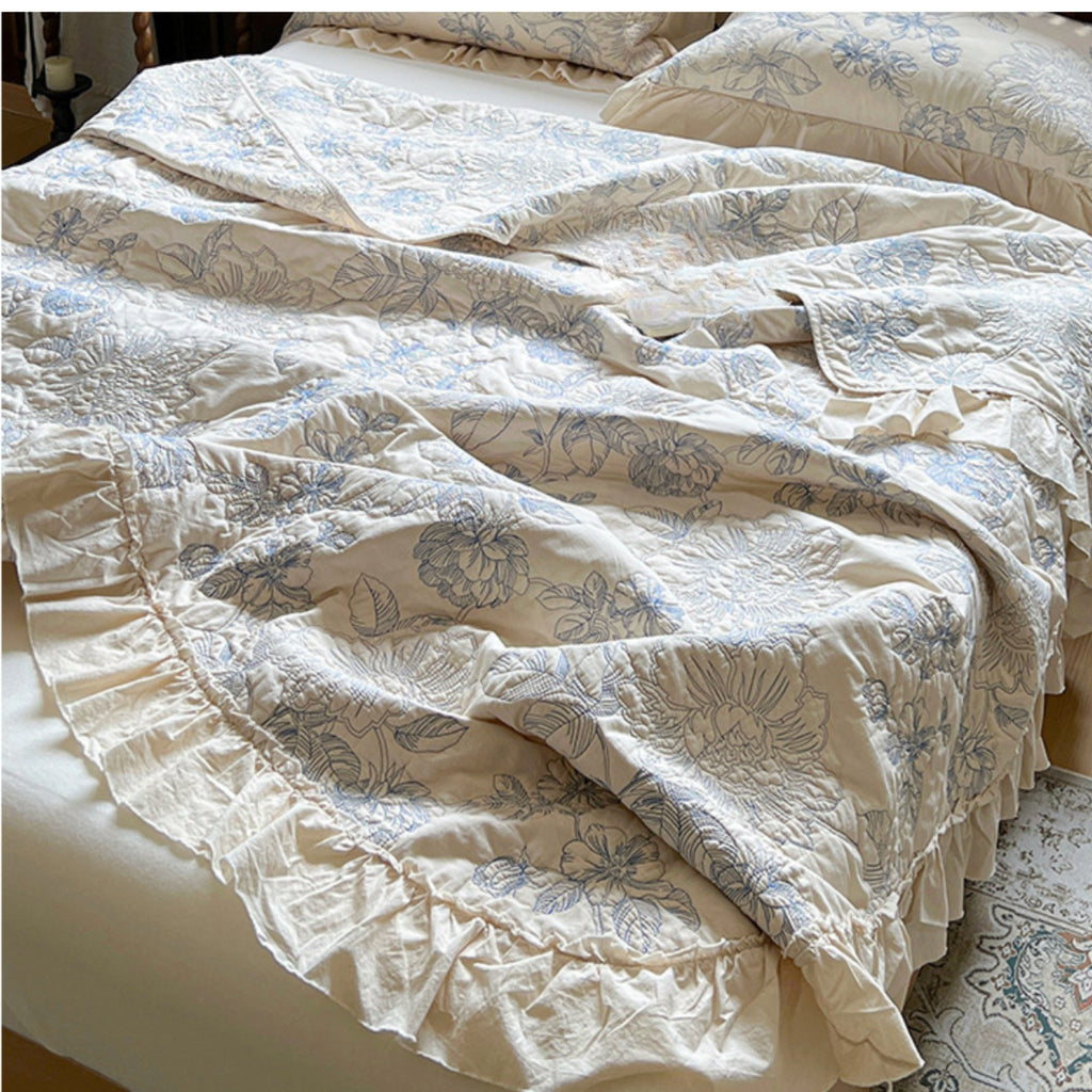 A close-up of a bed dressed in a cream-colored bedding set with a light blue floral pattern and ruffled trim, with matching pillows visible in the background.