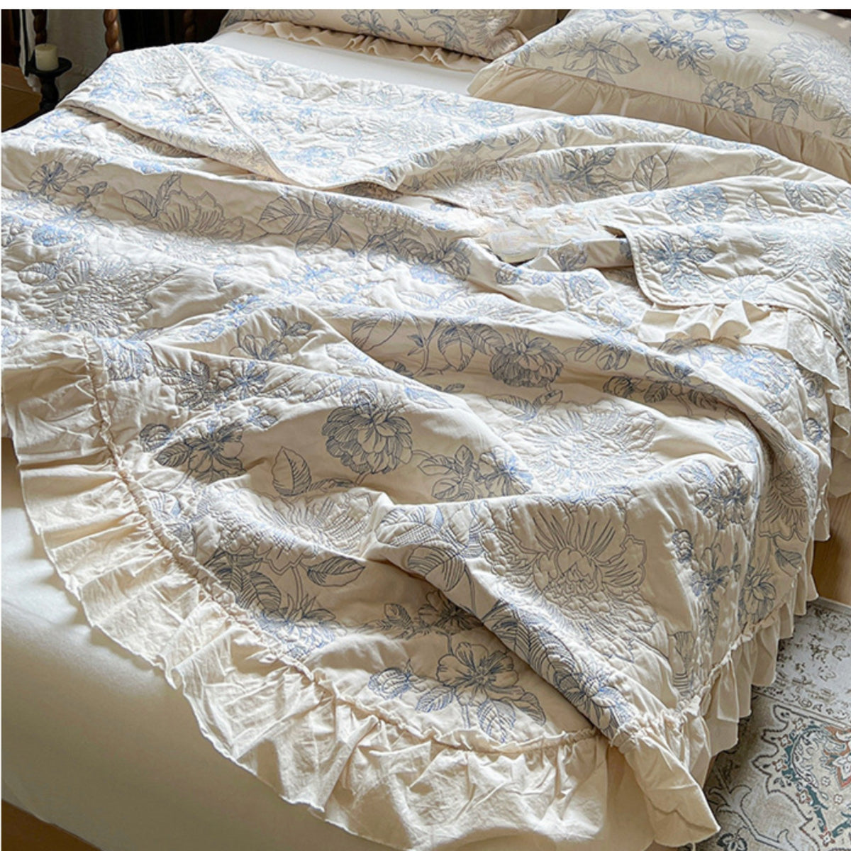 A close-up of a bed dressed in a cream-colored bedding set with a light blue floral pattern and ruffled trim, with matching pillows visible in the background.