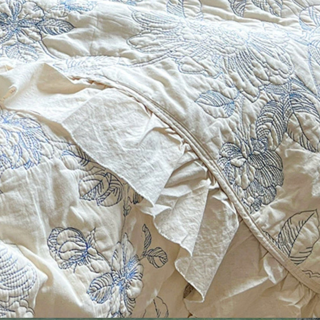 Close-up of a quilted textile in warm off-white/cream with blue floral embroidery and ruffled trim.