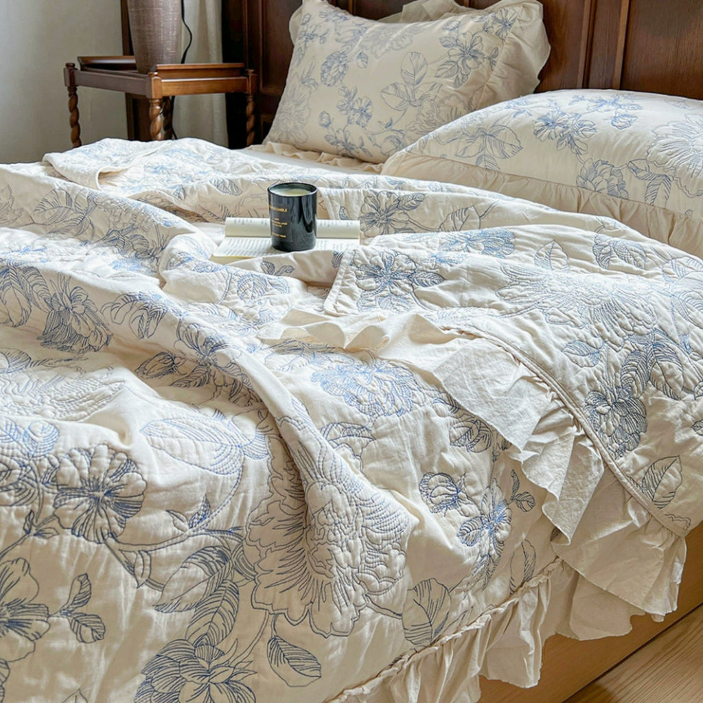 A close-up of a bed dressed in an off-white quilted bedding set with a blue floral pattern, including a comforter, matching pillow shams, and ruffled bed skirt, styled in a bedroom setting.