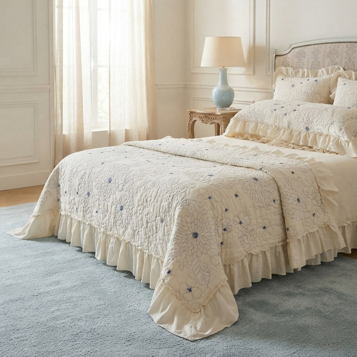 An elegant bed dressed in a cream-colored quilted bedding set with ruffled edges and delicate blue floral embroidery, styled in a bright, classic bedroom.