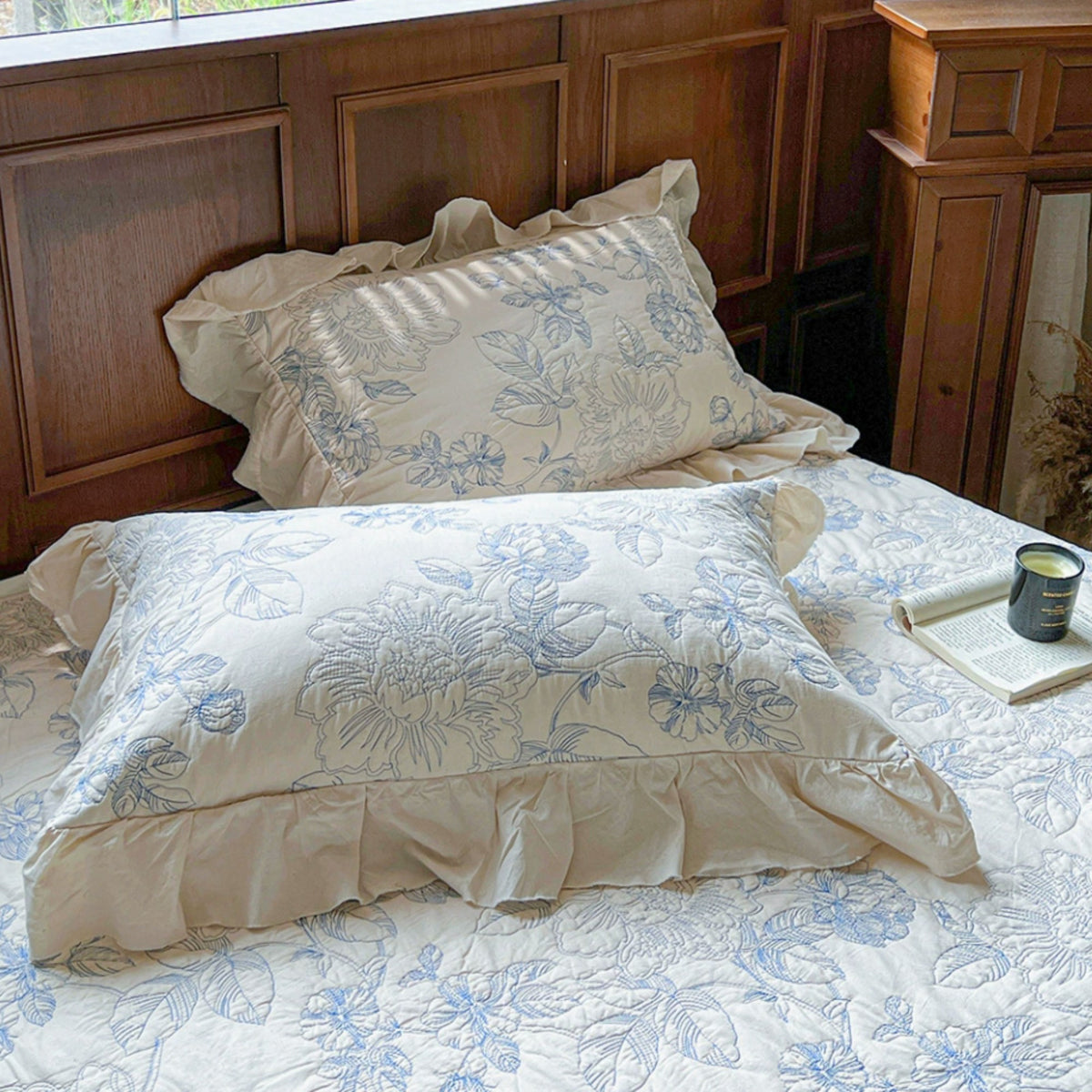 A bed dressed in cream bedding with a light blue floral quilt and matching pillow shams, styled in a warm bedroom setting with a wooden headboard, window light, and an open book on the bedside table.