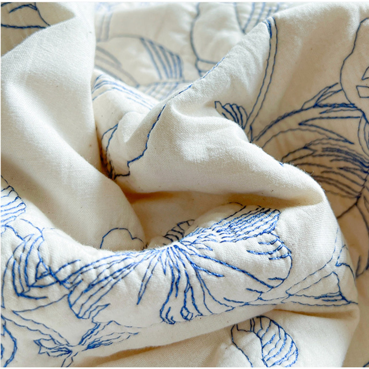 A close-up of a cream-colored quilted fabric with blue hand-stitched floral and leaf patterns.