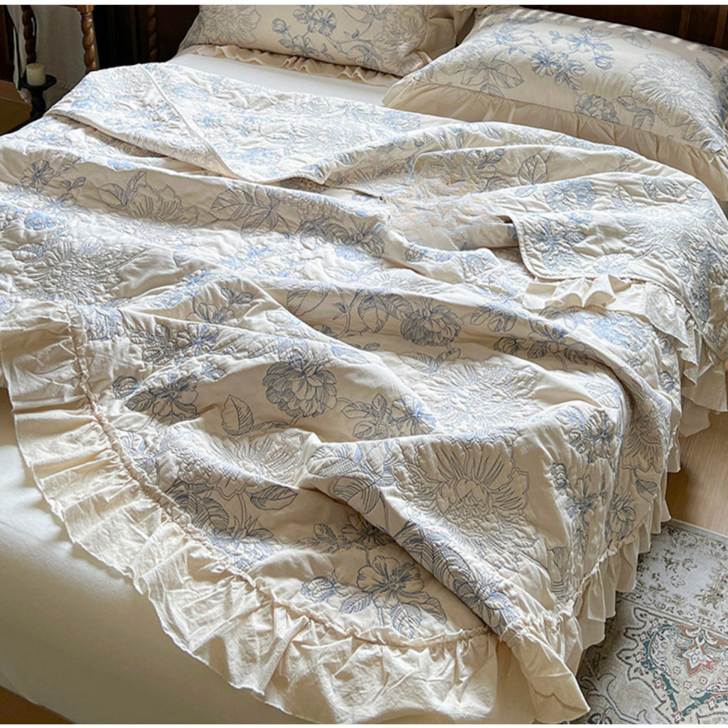A bed dressed in an off-white or cream bedding set with a dusty blue floral pattern and ruffled trim, shown in a bedroom setting.