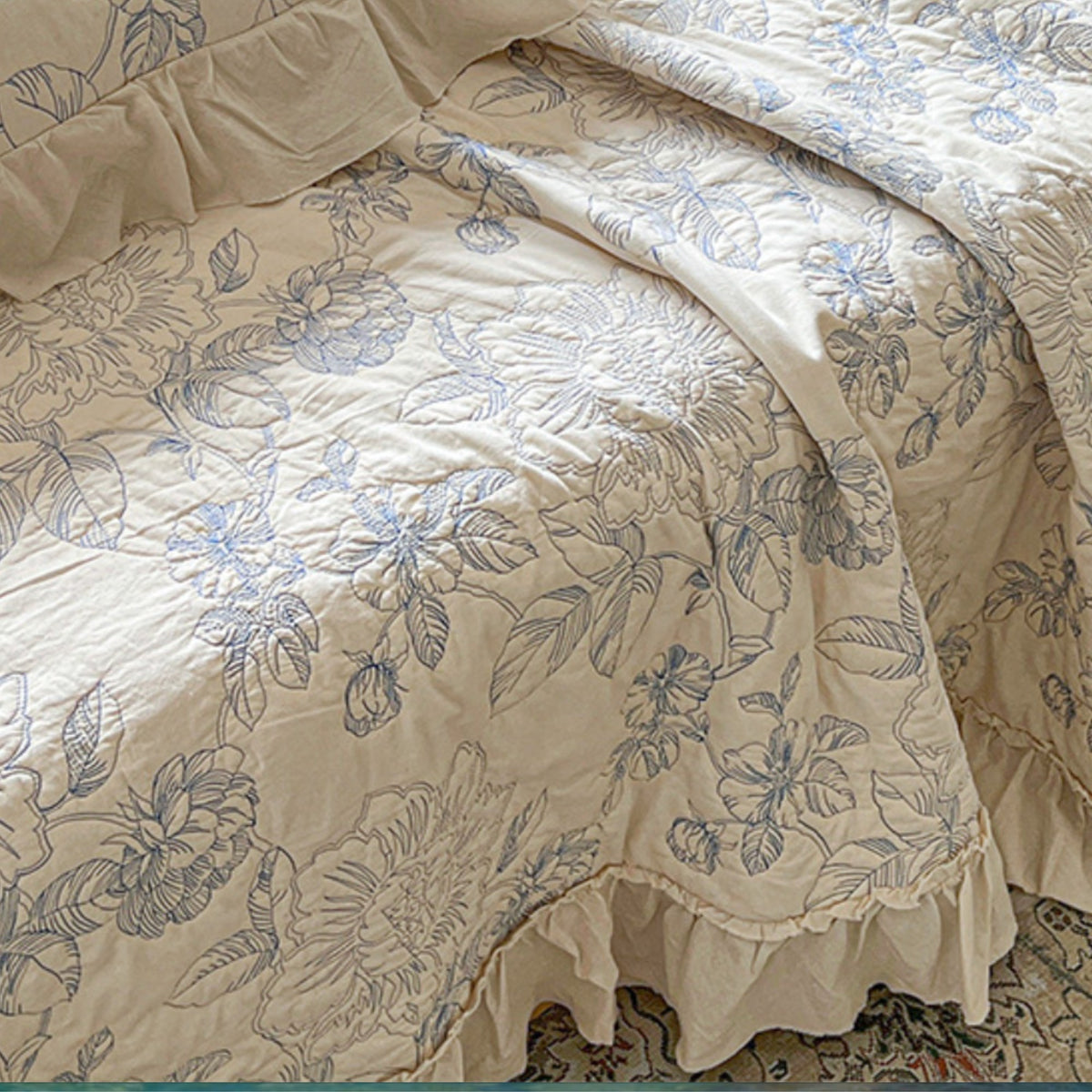 Beige quilted bedding set with a blue floral and leaf pattern, featuring a ruffled edge and matching pillowcases.