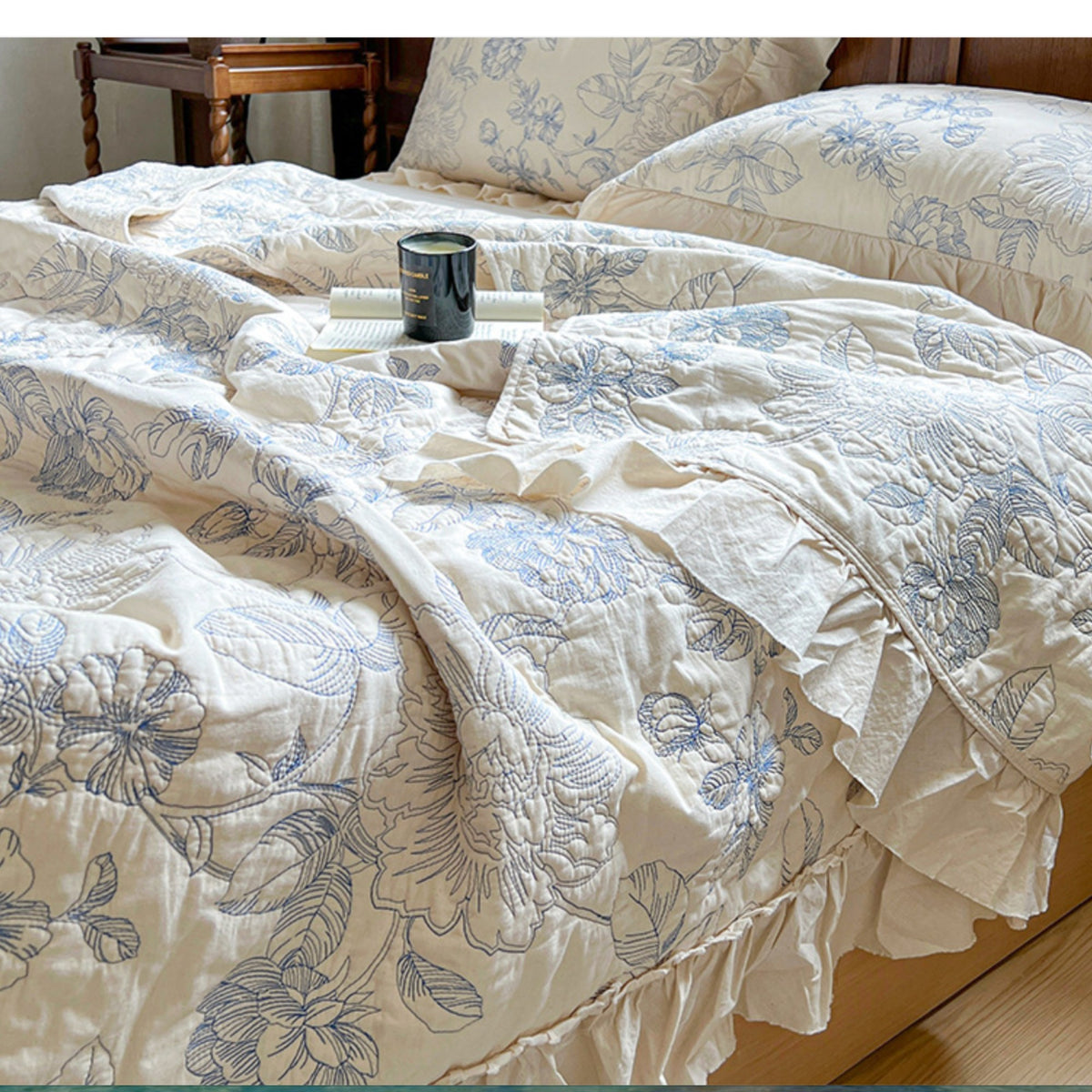 A close-up of a bed dressed in a cream-colored quilted coverlet and matching pillowcases, both featuring a blue floral and leaf pattern. The scene is styled with a small black candle and a white book on a tray, with a wooden headboard and side table visible in the background.