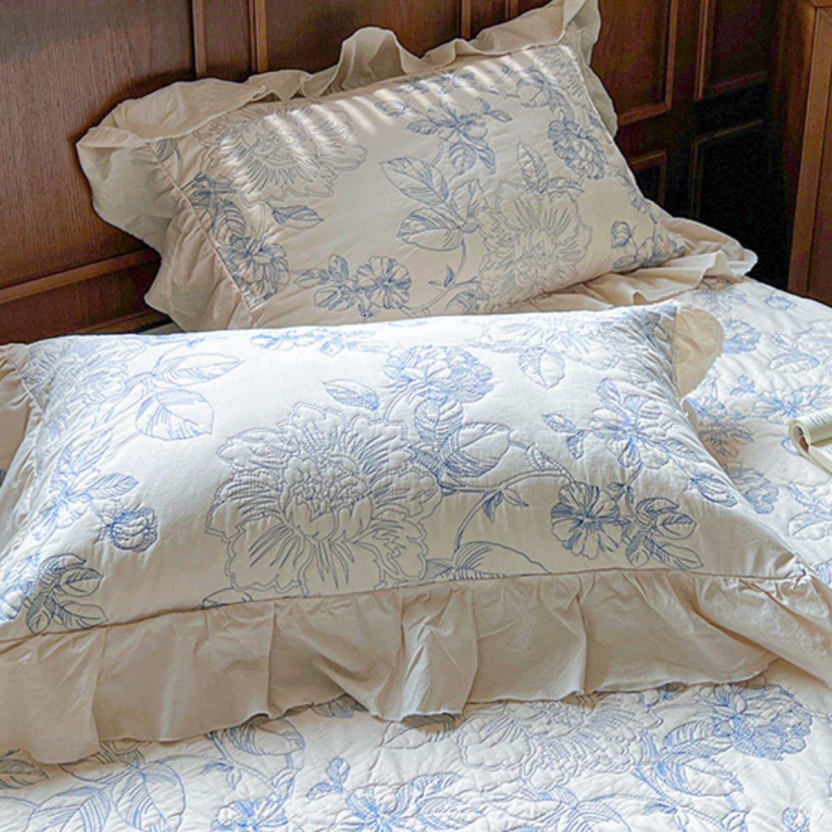 A close-up of a bed with cream-colored pillows and bedding featuring a light blue floral pattern, set against a dark wood headboard.