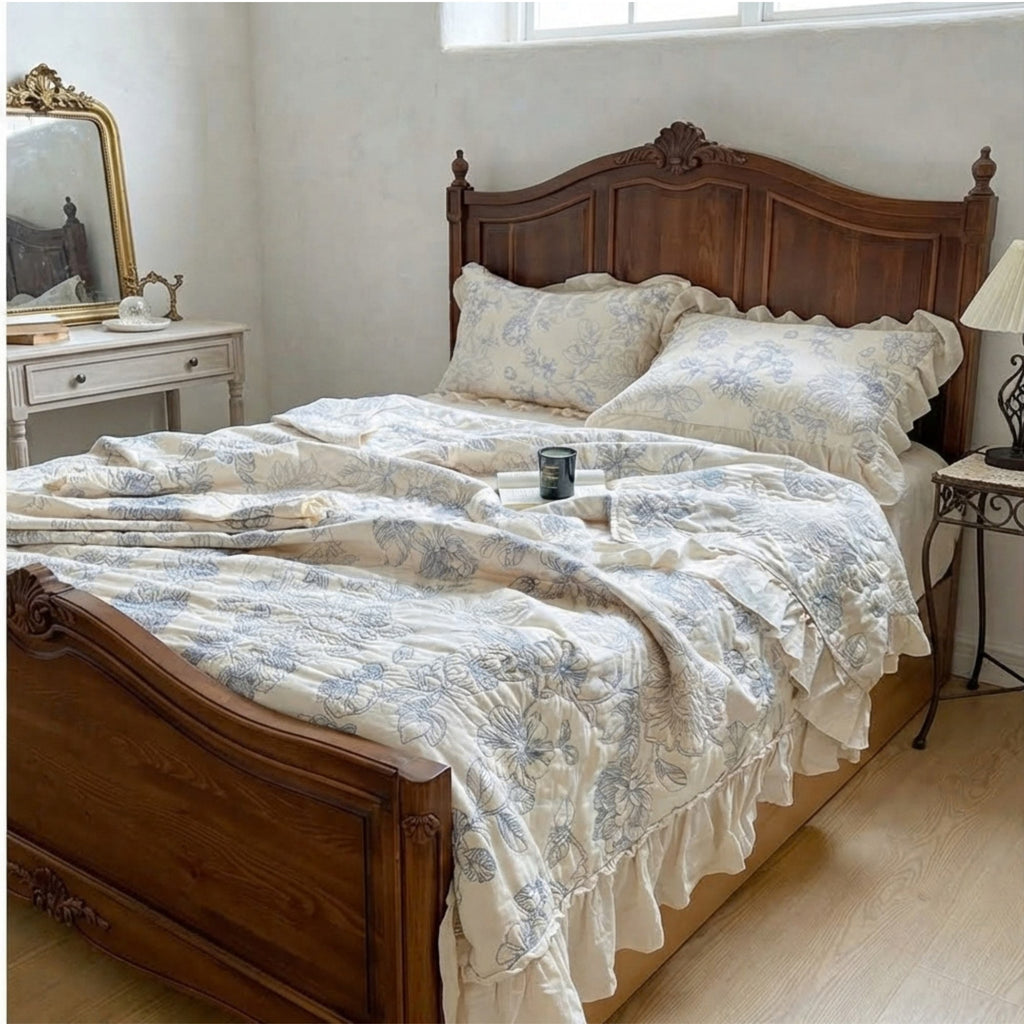 A rustic wooden bed frame dressed with a cream-colored  embroidered ruffled quilt and matching pillow shams featuring a blue floral toile pattern, set in a vintage-style bedroom.