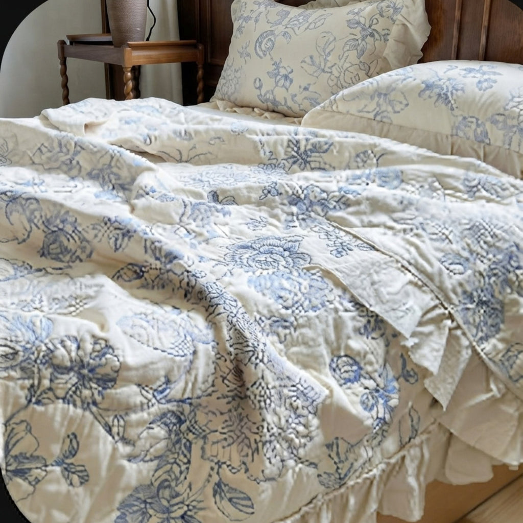 A close-up of a bed dressed in a cream-colored quilted comforter and matching pillow shams, both featuring a blue floral toile-style pattern. The bed is styled with a dark wood headboard and a matching wooden bedside table.