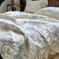 A close-up of a bed dressed in a cream-colored quilted comforter and matching pillow shams, both featuring a blue floral toile-style pattern. The bed is styled with a dark wood headboard and a matching wooden bedside table.