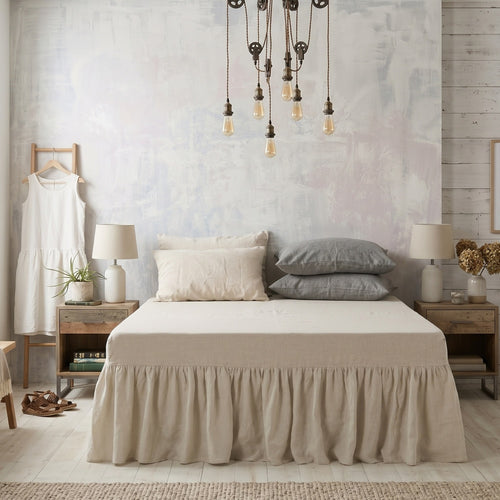 A neatly made bed in a styled bedroom, featuring a beige ruffled bed skirt, cream and gray pillows, matching bedside tables, lamps, and a rustic textured wall.