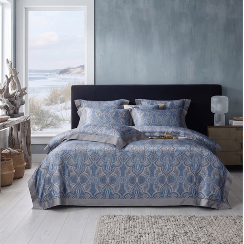 A bed dressed in a blue and gray ornate patterned bedding set with matching pillows, styled in a modern bedroom with a dark headboard, textured blue-gray wall, window view, and bedside table.