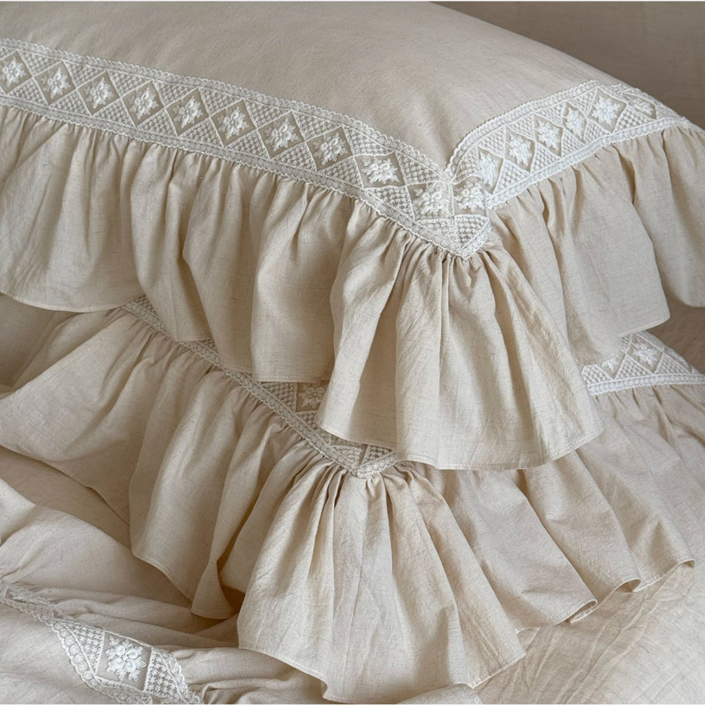 Beige ruffled bedding set with white embroidered lace trim, featuring a pillowcase and matching folded sheets.