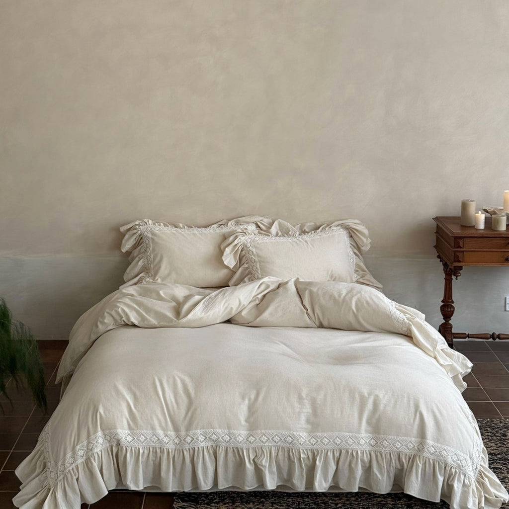 A bed dressed in soft off-white or cream bedding with ruffled edges and embroidered detailing along the bottom, styled in a rustic bedroom setting with a wooden side table and textured wall.
