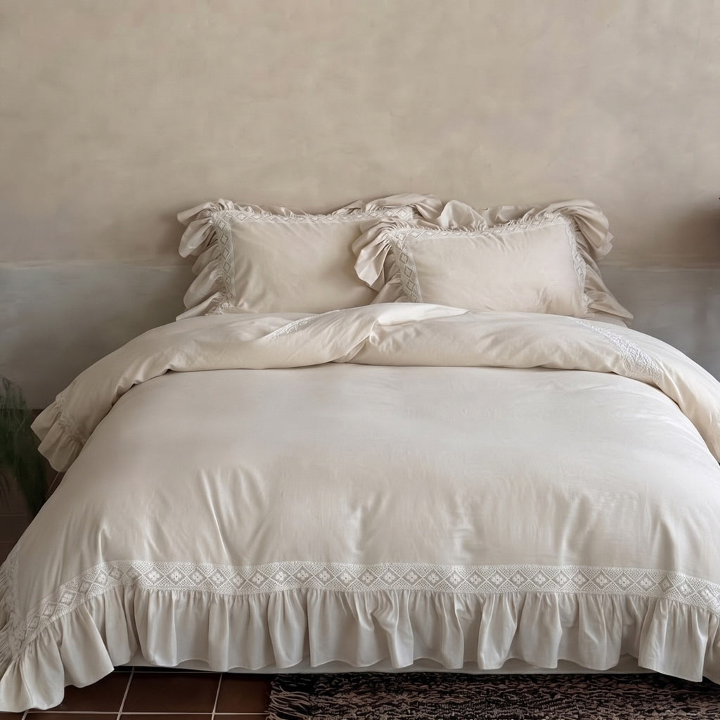 A bed dressed in a soft cream bedding set with ruffled pillows and a ruffled hem featuring embroidered geometric trim, set against a textured beige wall.
