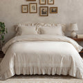 A neatly made bed dressed in a soft cream bedding set with ruffled pillows, a ruffled skirted duvet, and embroidered trim near the hem, styled in a warm neutral bedroom with framed wall art, a lamp, and a plant.