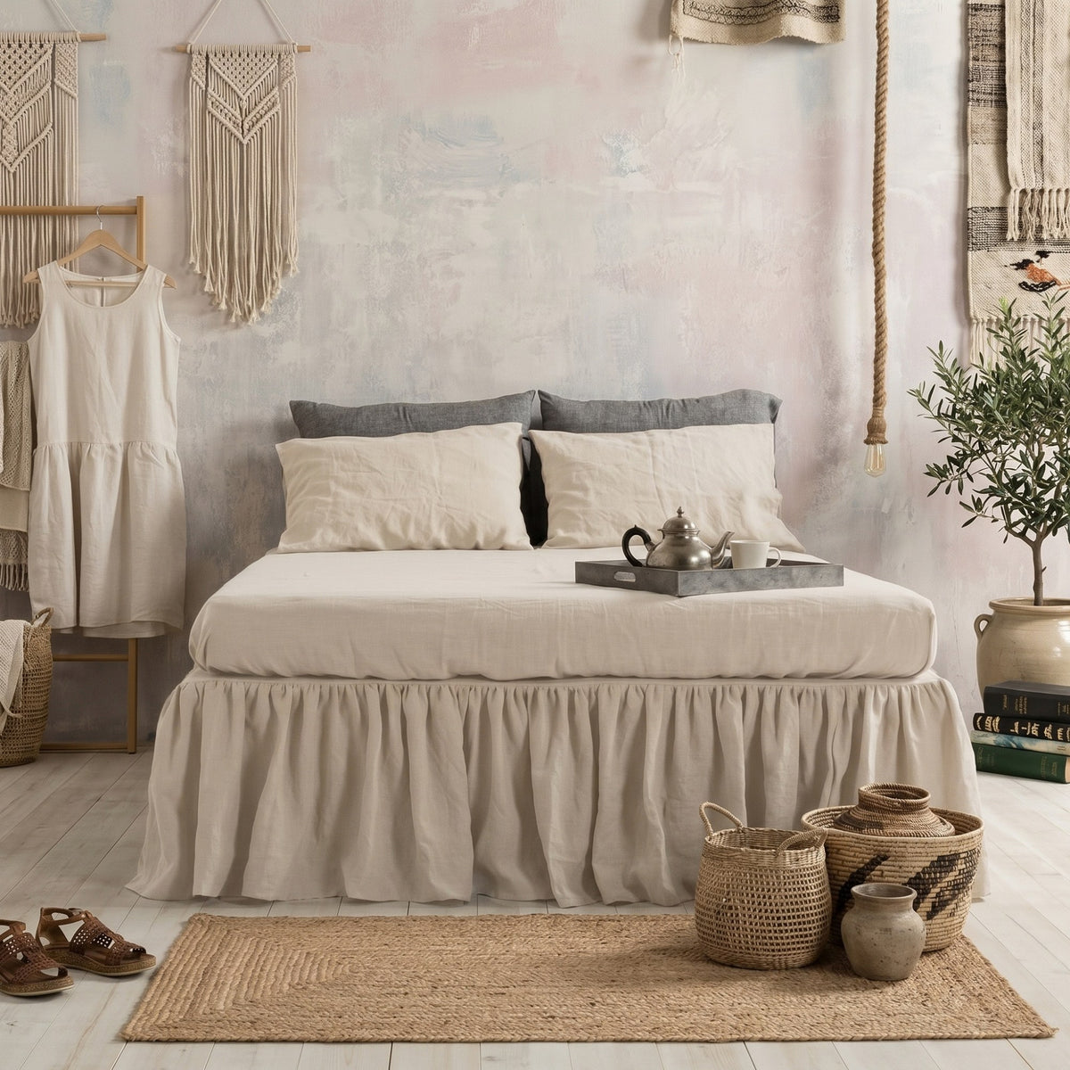 A bed dressed in a beige ruffled bed skirt with cream and gray pillows, styled in a boho bedroom with woven decor, a small dress on a hanger, sandals, baskets, and a plant.