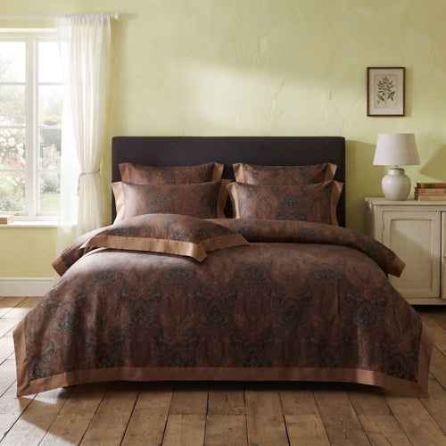 A bed dressed in a brown damask-patterned duvet cover and matching pillowcases with satin-like trim, styled in a bedroom with a pale yellow-green wall, wooden floor, window, and bedside table.