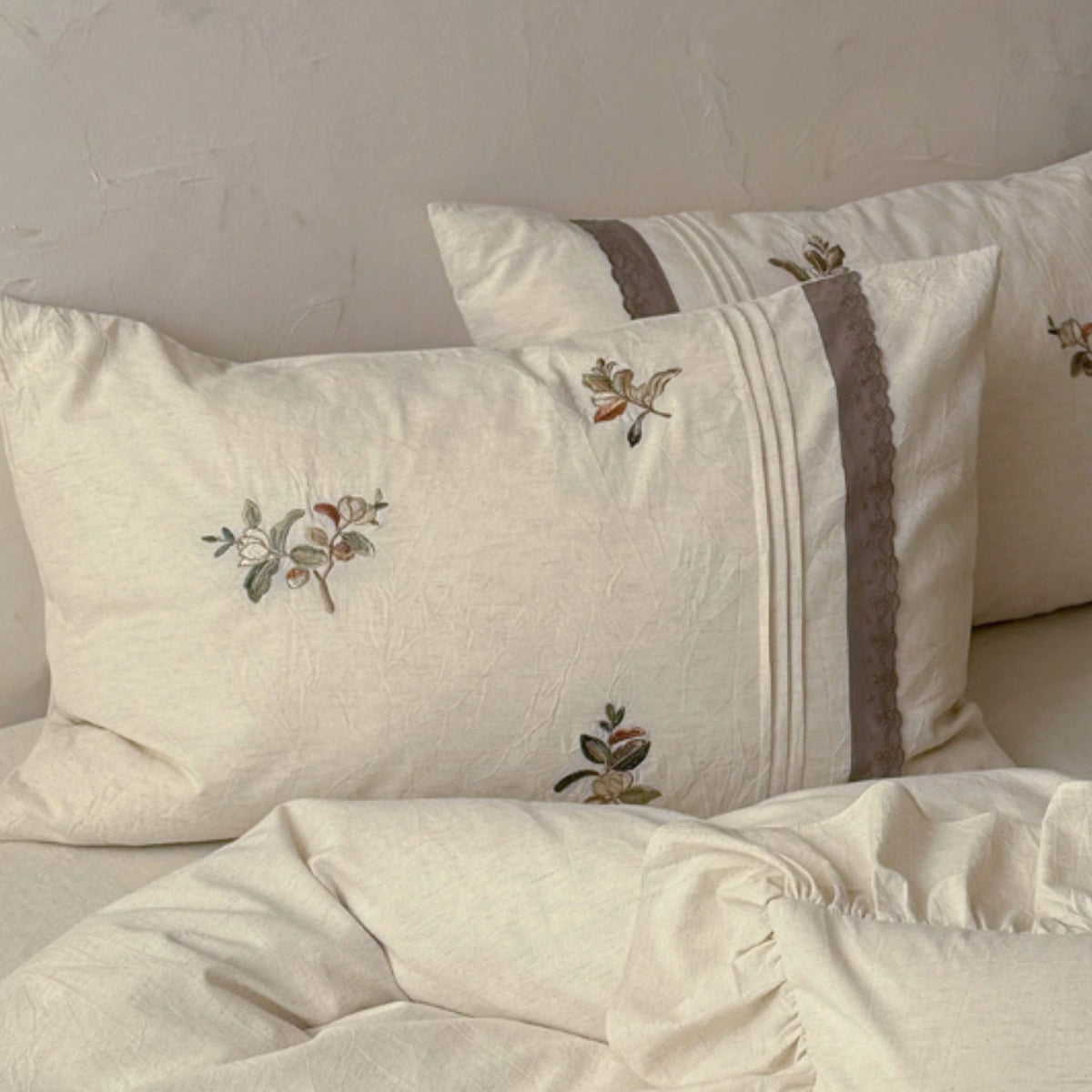 Cream-colored linen bedding set with embroidered floral motifs and a brown decorative stripe on the pillowcases, styled on a bed against a textured beige wall.