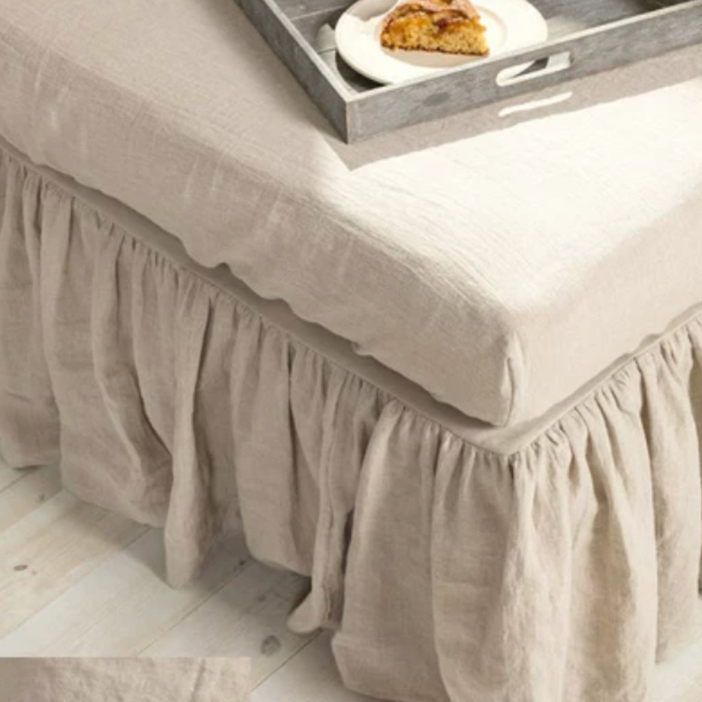 A beige linen bed skirt with a ruffled hemline draped over a bed, shown with a gray wooden serving tray on top containing a slice of cake.