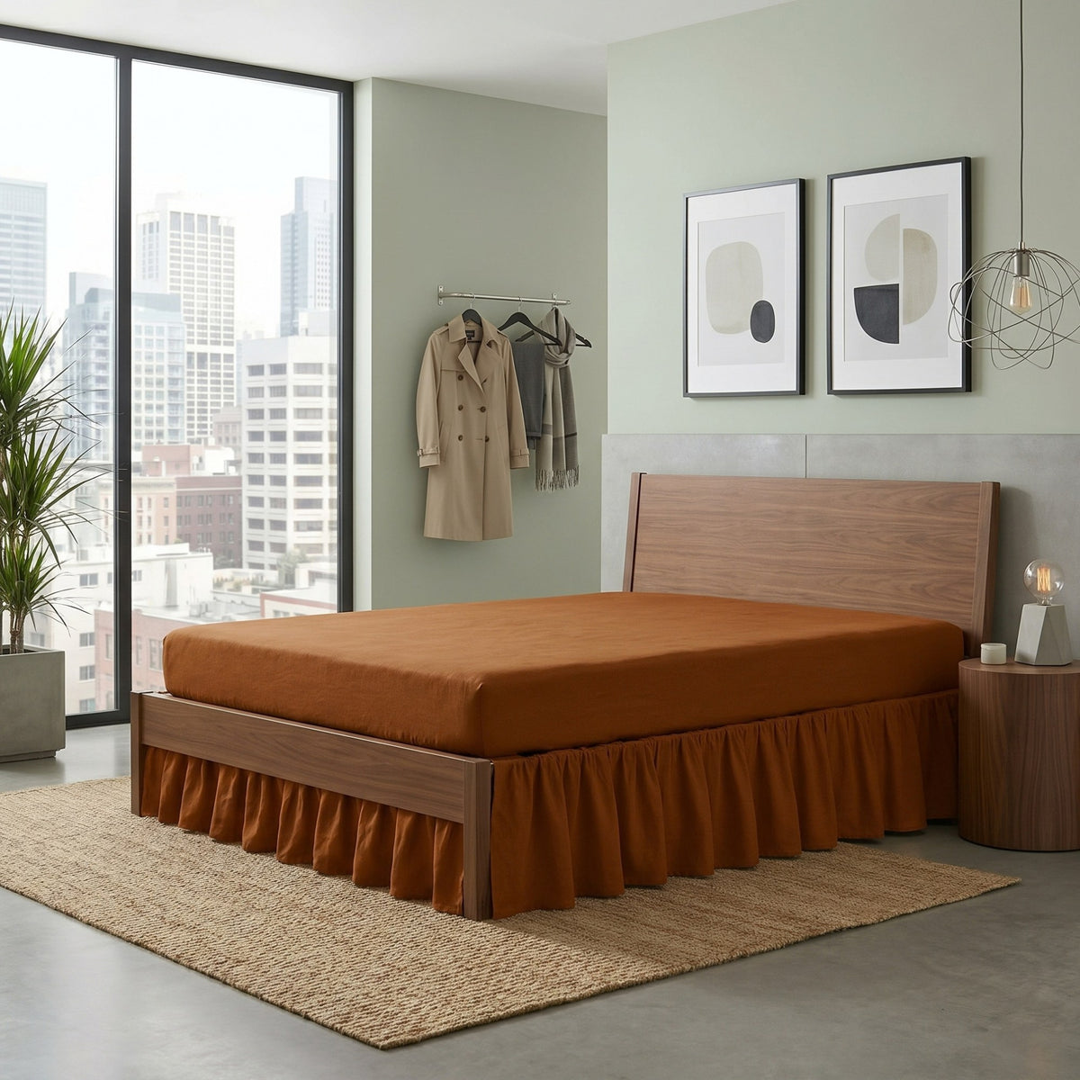 A modern platform bed with a medium brown wood frame and a rust-orange bed skirt, styled in a contemporary bedroom with a city view window, a woven rug, and minimalist wall art.