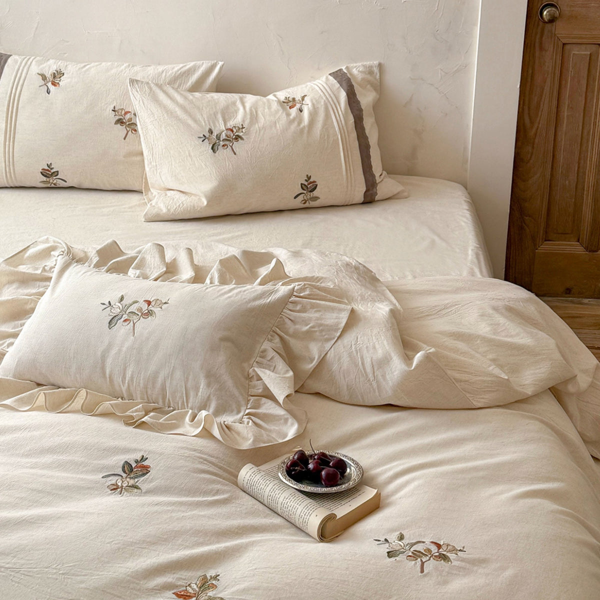 A neatly made bed with cream-colored bedding, including a duvet cover, pillows, and ruffled trim, decorated with small floral embroidery. A small book and a bowl of dark cherries sit on the bedding, with a wooden door in the background.