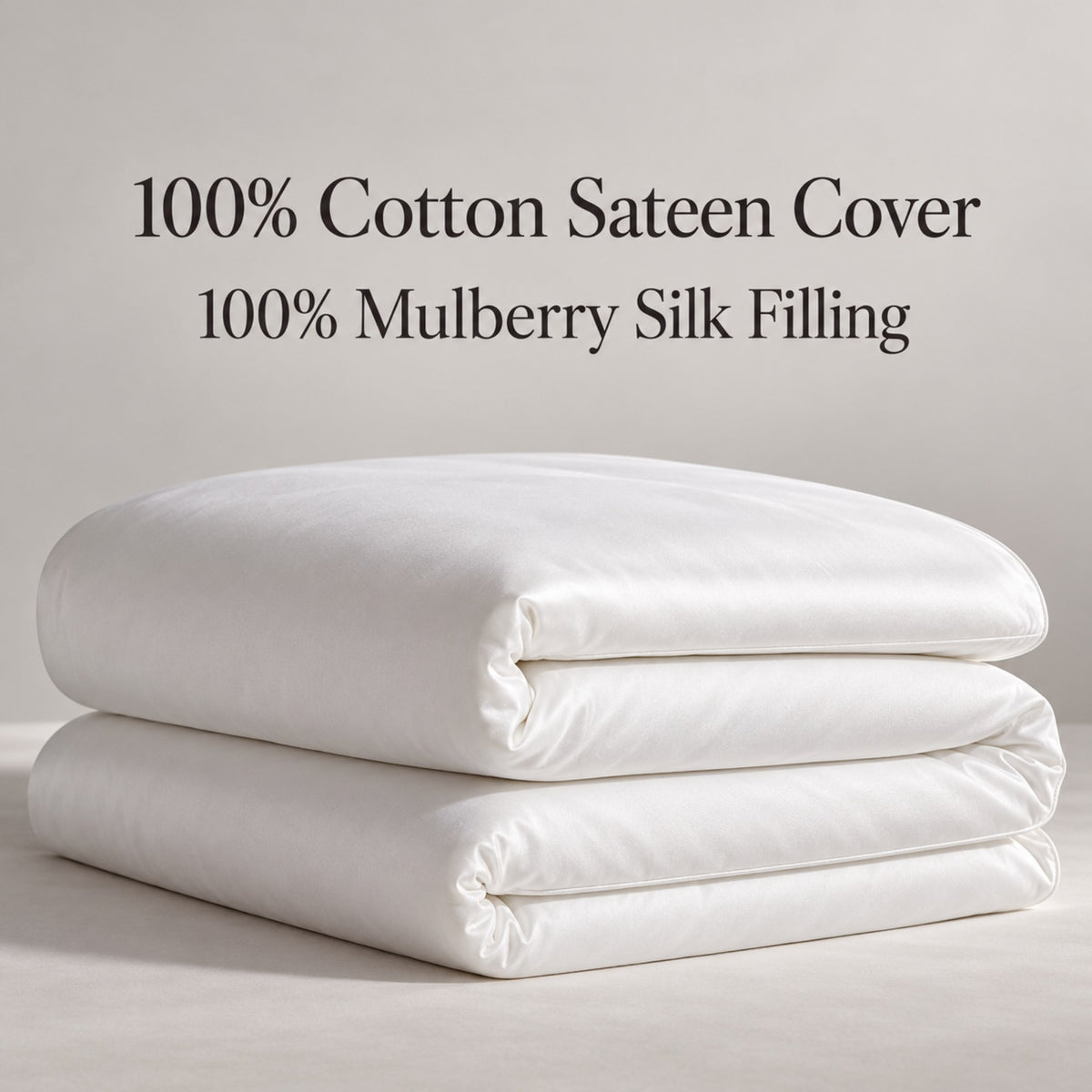 Two folded white bedding pieces stacked on a light neutral surface, with text above reading '100% Cotton Sateen Cover' and '100% Mulberry Silk Filling'.