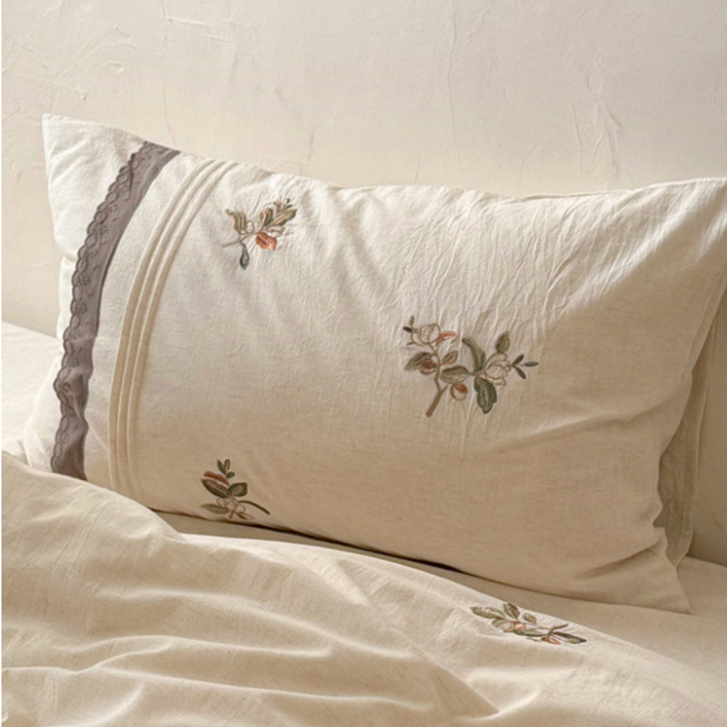 An off-white linen pillowcase with delicate floral embroidery and a gray lace trim, resting on a matching cream-colored bedsheet.