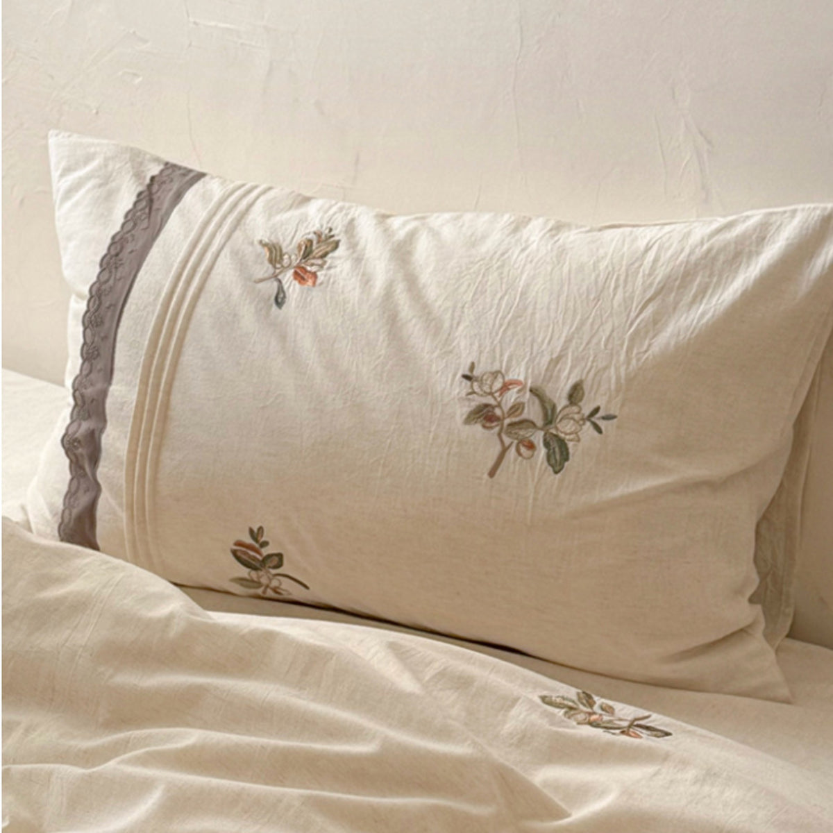 An off-white linen pillowcase with delicate floral embroidery and a gray lace trim, resting on a matching cream-colored bedsheet.